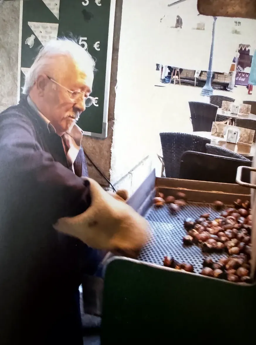 En Paco garbellant les castanyes acabades de torrar, amb els preus a la pissarra del darrere
